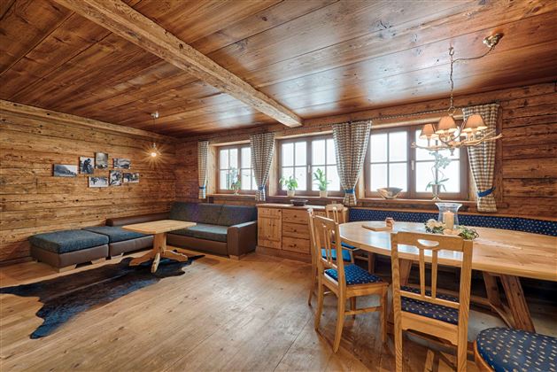 Ein gemütlicher Wohnraum mit Holzwänden und einem großen Esstisch. Große Fenster lassen viel Licht herein und bieten einen Blick nach draußen.