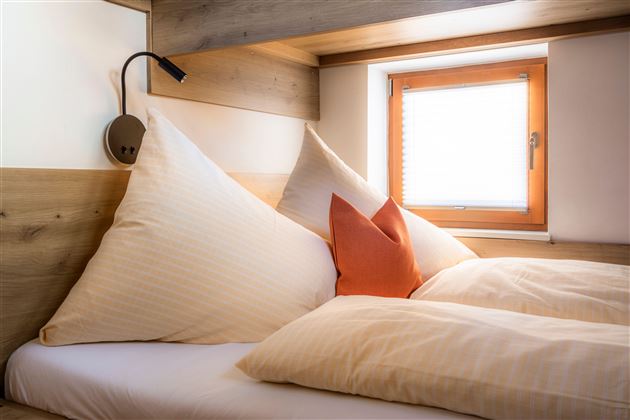 A cozy bedroom with large, pointed pillows and an orange decorative cushion. The window lets in natural light and creates an inviting atmosphere.