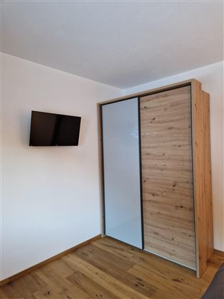 A simple room with a TV on the wall and a modern wardrobe with sliding doors. The floor is made of wood and the ambiance is bright and welcoming.