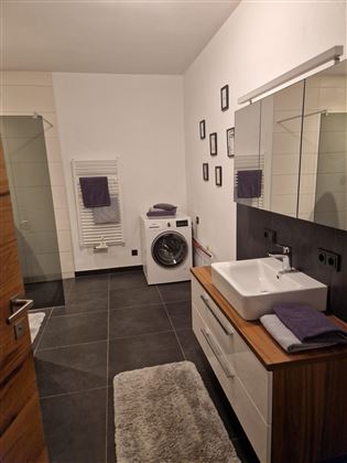 A modern bathroom with a washing machine and a large mirror. The walls are light, and there are towels in purple tones.