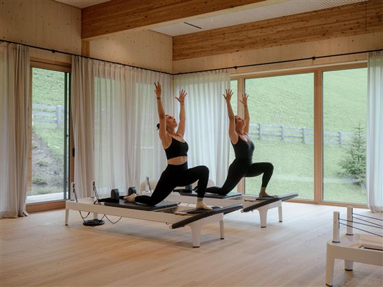 Two women are doing Pilates in a modern room with plenty of natural light. In the background, there are windows overlooking a green landscape.