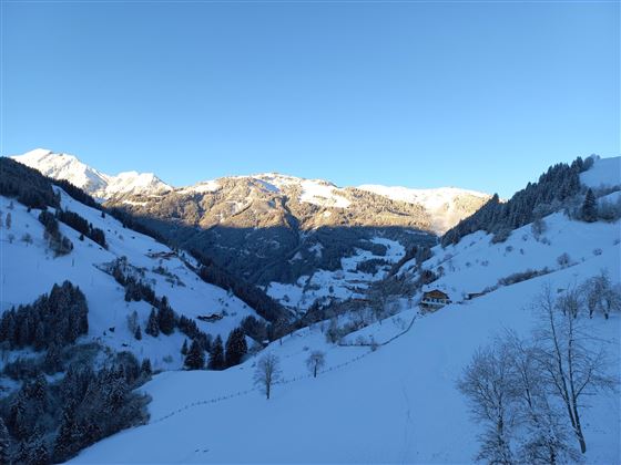 A snowy mountain landscape with high mountains and a clear sky. The valley is serene and picturesque with scattered trees and a small house.