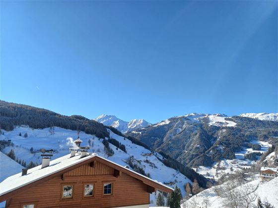 A picturesque mountain landscape with snow-covered hills and a clear blue sky. In the foreground stands a wooden house.