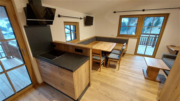 A modern kitchen corner with wooden shelves and a dining table. Large windows provide a view of the landscape.