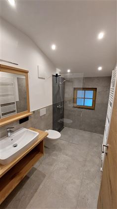 A modern bathroom with a shower and a wooden sink. It has a bright, neutral color scheme and a window for natural light.