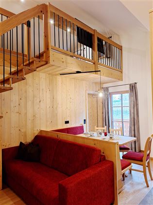 A cozy apartment with wood paneling, a red sofa, and a dining area. In the upper area, there is a balcony overlooking the room.