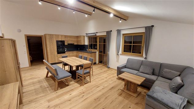 A modern kitchen with a large dining table and a cozy living room. Bright wood surfaces and soft lighting create a warm atmosphere.