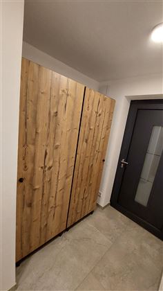 Ein Holzschrank mit einer natürlichen, strukturierten Oberfläche steht in einem Flur. Die Tür des Flurs ist dunkel und hat ein großes Fenster.