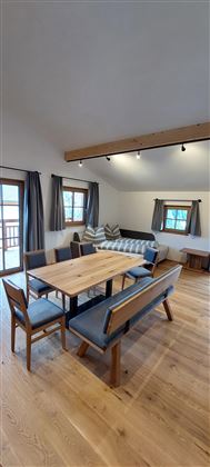 Ein gemütlicher Wohnraum mit einem Holztisch und Sitzbänken. Große Fenster lassen viel Licht herein.