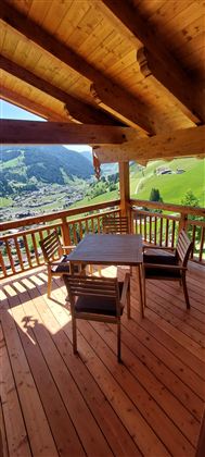 Eine Holzterrasse mit einem Tisch und Stühlen bietet einen schönen Blick auf die grüne Landschaft. Der sonnige Himmel und die Berge im Hintergrund schaffen eine einladende Atmosphäre.