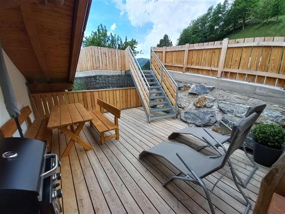 A cozy wooden terrace with a grill, a table, and lounge chairs. In the background, there is a staircase and a view of the green countryside.