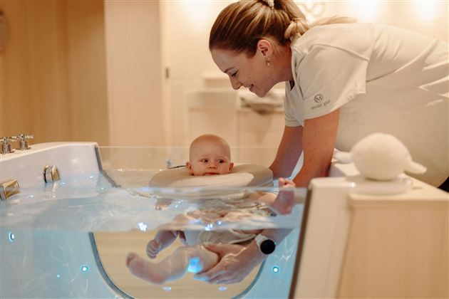 A woman is standing above a baby in a special bathtub area. The baby is safely sitting in the water and smiling, while the woman looks at it kindly.