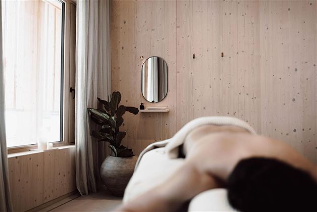 A relaxing spa room with wooden paneling and plenty of natural light. In the foreground, a person is lying on a lounge chair, surrounded by plants.