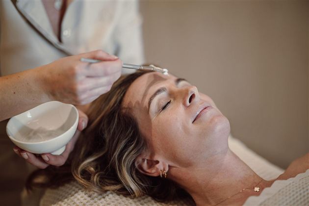 A woman is receiving a facial treatment in a relaxing environment. A hand is applying cream to her forehead.