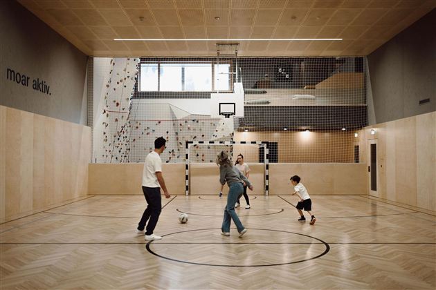 An interior space with a sports field where four people are playing soccer. The walls are decorated with climbing elements and large windows provide light.