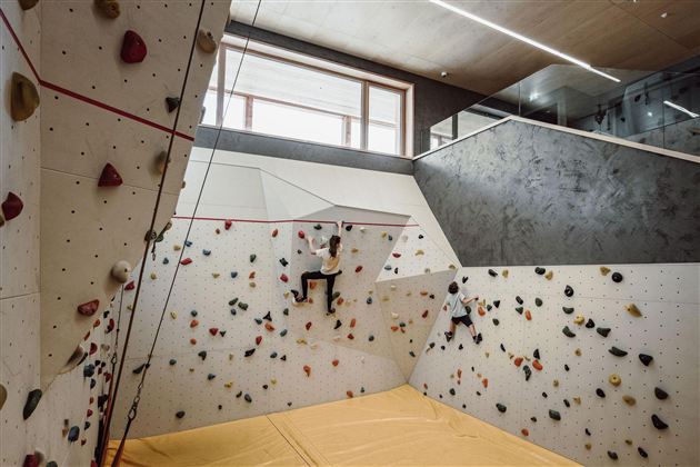 A modern climbing wall with various grips in a bright indoor space. One person is climbing on the wall, while other areas of the room are visible.