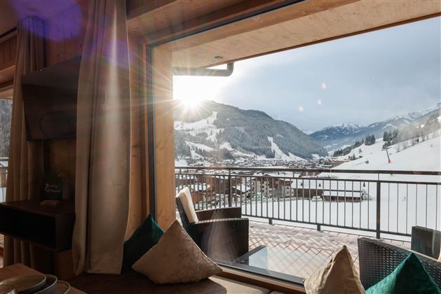 Ein gemütlicher Raum mit großen Fenstern, die auf schneebedeckte Berge und ein sonniges Tal blicken. Drapierte Vorhänge und bequeme Kissen schaffen eine einladende Atmosphäre.
