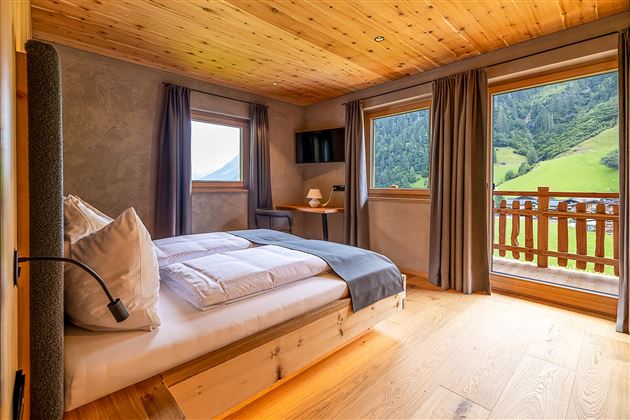 Ein gemütliches Schlafzimmer mit Holzdecke und großen Fenstern. Der Blick zeigt eine grüne Landschaft mit Bergen und Blick auf den Balkon.