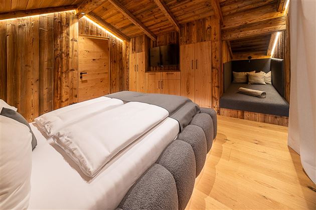 Ein gemütliches Schlafzimmer mit Holzwänden und einem modernen Bett. Im Hintergrund befindet sich eine sitzecke mit Kissen.