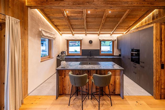 Eine moderne Küche mit Holzdecken und eleganten, grauen Schränken. Eine gemütliche Bar mit zwei Stühlen ergänzt den offenen Raum.