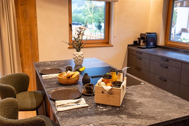 Eine moderne Küche mit einem Stein Tisch und stilvollen Stühlen. Auf dem Tisch stehen Frühstücksartikel in einem Korb sowie frische Früchte und Getränke.
