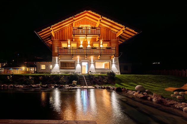Ein wunderschönes Holzhaus bei Nacht, beleuchtet von warmem Licht. Im Vordergrund spiegelt sich das Haus im ruhigen Wasser eines kleinen Teichs.