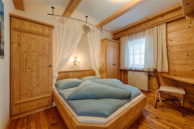 A cozy wooden bedroom with a large bed and blue bedding. The decor is rustic and inviting, featuring a window and a chair.