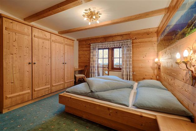 A cozy bedroom with wooden furniture and a large window. The bed is decorated with blue pillows.