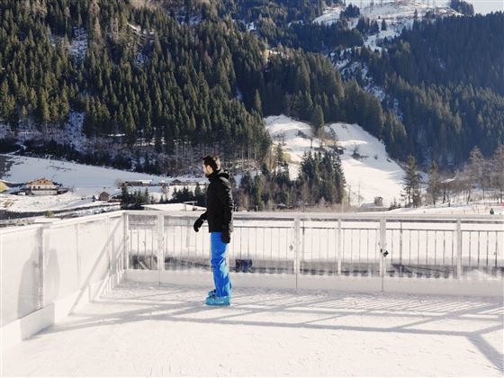 A man stands on an ice surface in a wintry landscape. Snow-covered mountains and forests can be seen in the background.