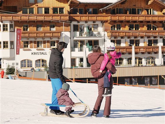 A family stands in the snow while pulling a sled. Beautiful mountain houses are visible in the background.