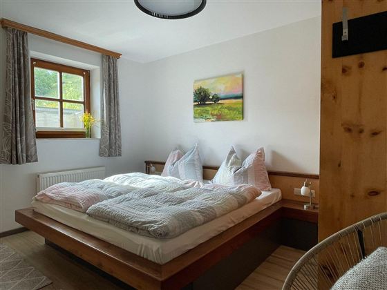 Ein gemütliches Schlafzimmer mit einem großen Bett und weichen Bettdecken. Ein Fenster lässt Tageslicht herein, und an der Wand hängt ein Landschaftsgemälde.