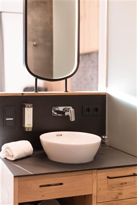 Ein modernes Badezimmer mit einem eleganten Waschbecken und einem spiegelnden Kosmetikspiegel. Die Holzdetails und die minimalistische Gestaltung schaffen eine einladende Atmosphäre.