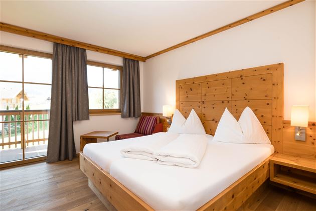 Ein gemütliches Schlafzimmer mit einem großen Doppelbett und Holzdetails. Die Fenster bringen viel Tageslicht und blicken auf einen Balkon.