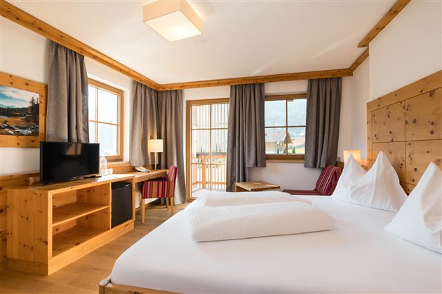 Ein gemütliches Zimmer mit einem Doppelbett und Holzmöbeln. Große Fenster lassen viel Licht herein und bieten einen Blick nach draußen.