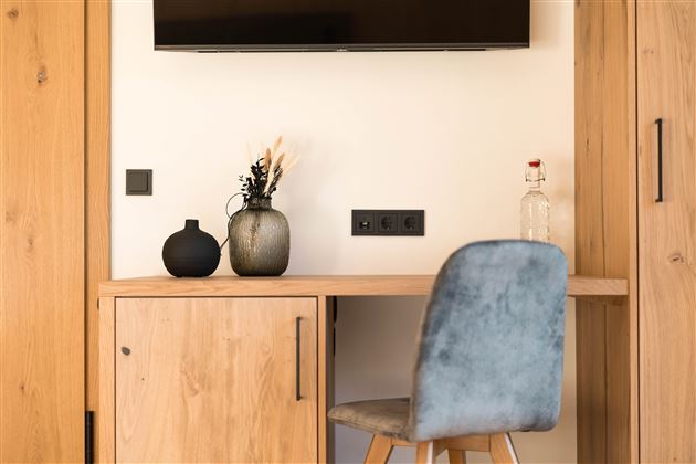 Ein gemütlicher Arbeitsplatz mit einem Holzschreibtisch und einem blauen Stuhl. Auf dem Tisch stehen eine Vase, ein schwarzes Gefäß und eine Flasche, während ein Fernseher an der Wand montiert ist.