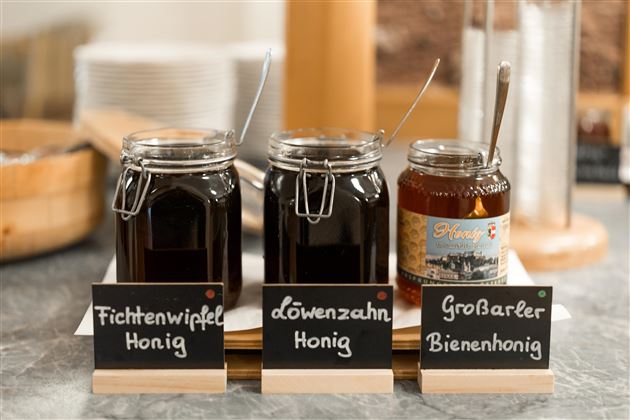 Drei Gläser mit verschiedenen Honigen stehen auf einem Tisch. Jedes Glas ist mit einem Schild gekennzeichnet: Fichtenwipfel-Honig, Löwenzahn-Honig und Großartler Bienenhonig.