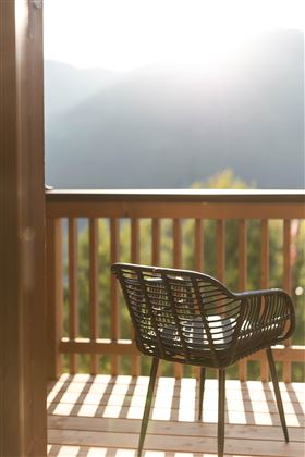 Ein stilvoller Stuhl steht auf einem Balkon mit Holzgeländer. Im Hintergrund sind sanfte Hügel und die Sonne scheint.