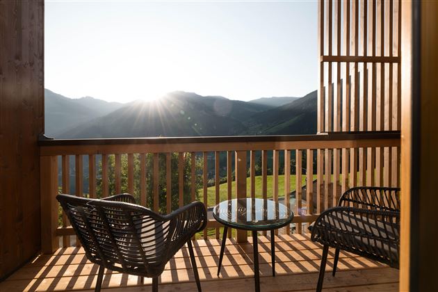 Ein Balkon mit zwei gemütlichen Stühlen und einem Tisch. Im Hintergrund sind die Berge und die aufgehende Sonne zu sehen.