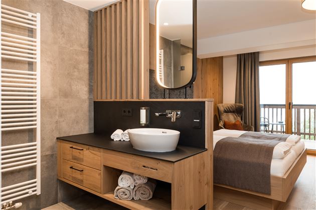 Ein modernes Badezimmer mit einem eleganten Waschbecken und Holzdetails. Im Hintergrund ist ein gemütliches Schlafzimmer mit Fenster und einem Balkon sichtbar.