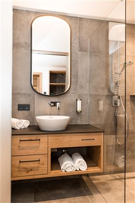 Ein modernes Badezimmer mit einer gläsernen Dusche und einem runden Spiegel. Auf der Holztheke liegen frische Handtücher bereit.