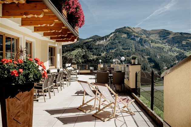 Eine sonnige Terrasse mit gemütlichen Liegestühlen und blühenden Blumen. Im Hintergrund sind die beeindruckenden Berge sichtbar.