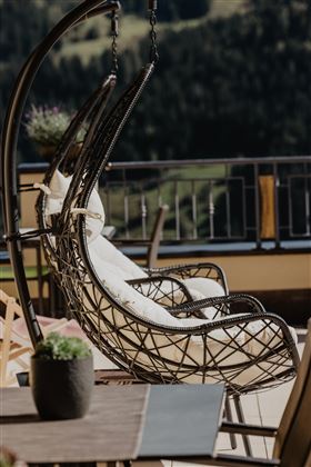 Ein stilvoller Hängesessel mit weißen Kissen steht auf einer Terrasse. Im Hintergrund sind grüne Berge und eine friedliche Landschaft zu sehen.
