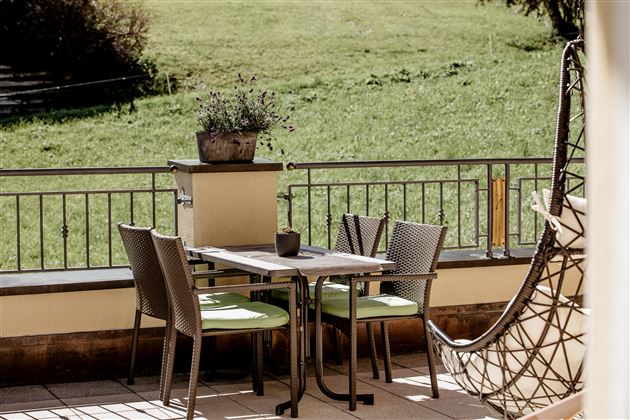 Ein gemütlicher Balkon mit einem Tisch und Stühlen. Im Hintergrund sieht man eine grüne Wiese und einen hängenden Stuhl.