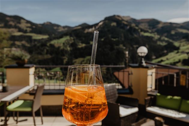 Ein Glas mit einem orangefarbenen Getränk steht auf einer Terrasse mit Blick auf grüne Berge. Die Atmosphäre ist entspannt und einladend.