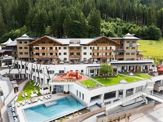 A luxurious hotel building surrounded by trees and green landscape. In the foreground, there is a pool area with loungers and a well-kept garden area.