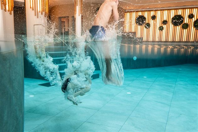 An indoor pool with two people jumping into the water. Splashes and waves occur as they dive into the cool water.