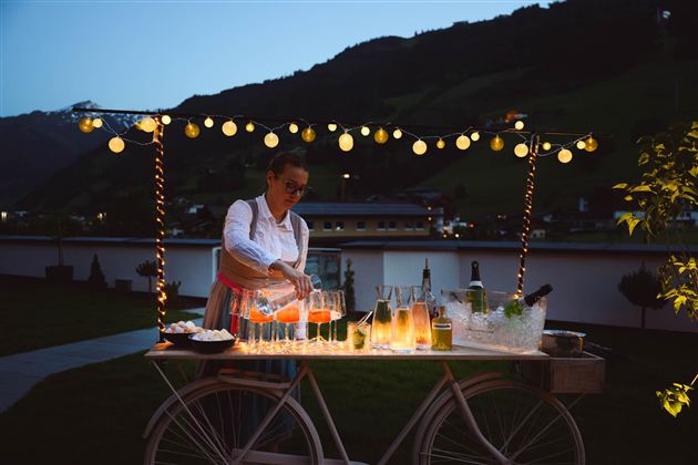 A woman is preparing drinks at a decorated cart at night. The surroundings are adorned with lights and offer a beautiful view of the landscape.