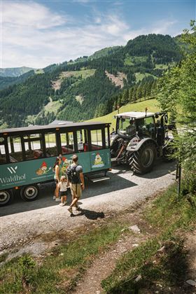 A family is standing by a path in front of a tractor and a cart in a picturesque mountainous landscape. In the background, green hills and a clear blue sky can be seen.