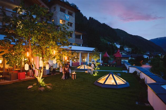 A beautiful garden with illuminated pavilions and an inviting atmosphere. In the background, mountains and a clear evening sky can be seen.