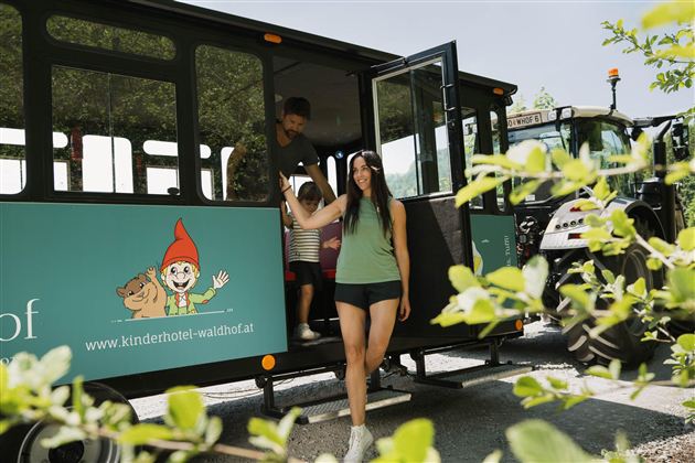 A family is getting off a bus with a cheerful design. In the background, a tractor and a garden landscape can be seen.